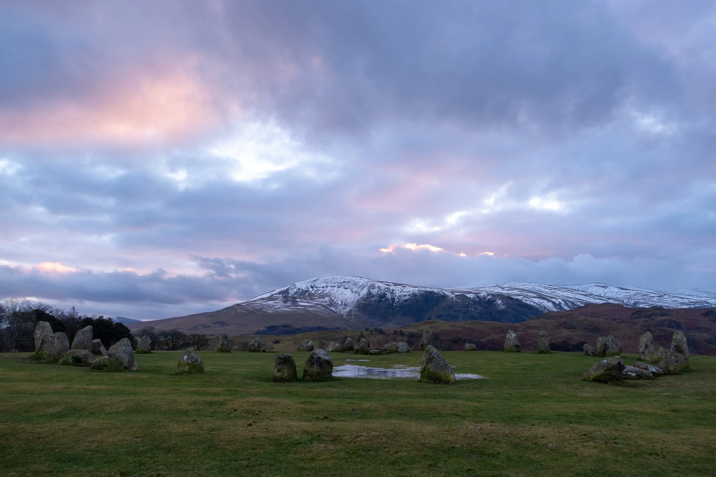 Castlerigg Stone Circle, in a grassy field with a mountain in the background, partly snow-capped, under a cloudy pink and grey sky.