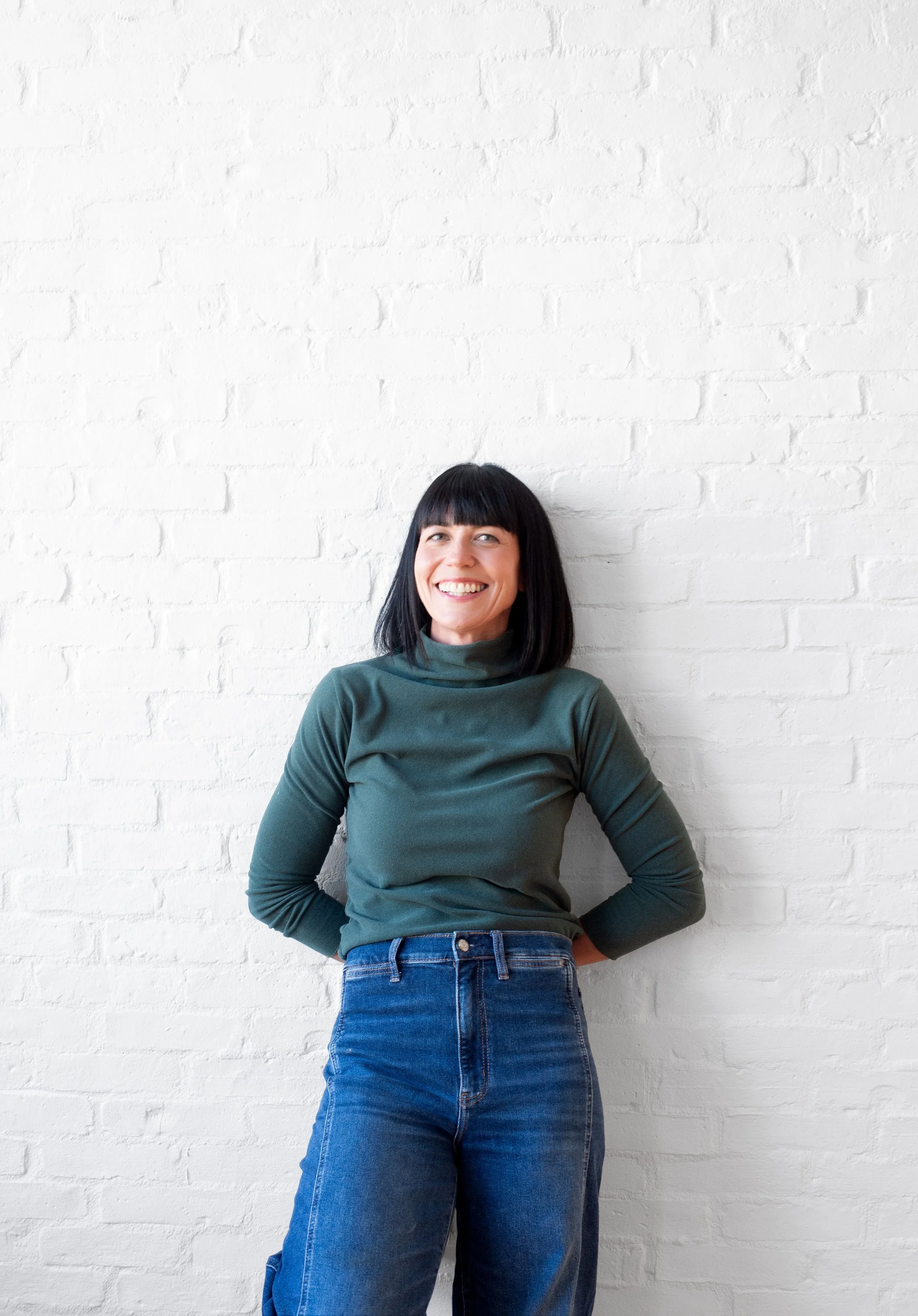 A woman with black hair and bangs smiling, wearing a green turtleneck sweater and blue jeans, standing against a white brick wall.