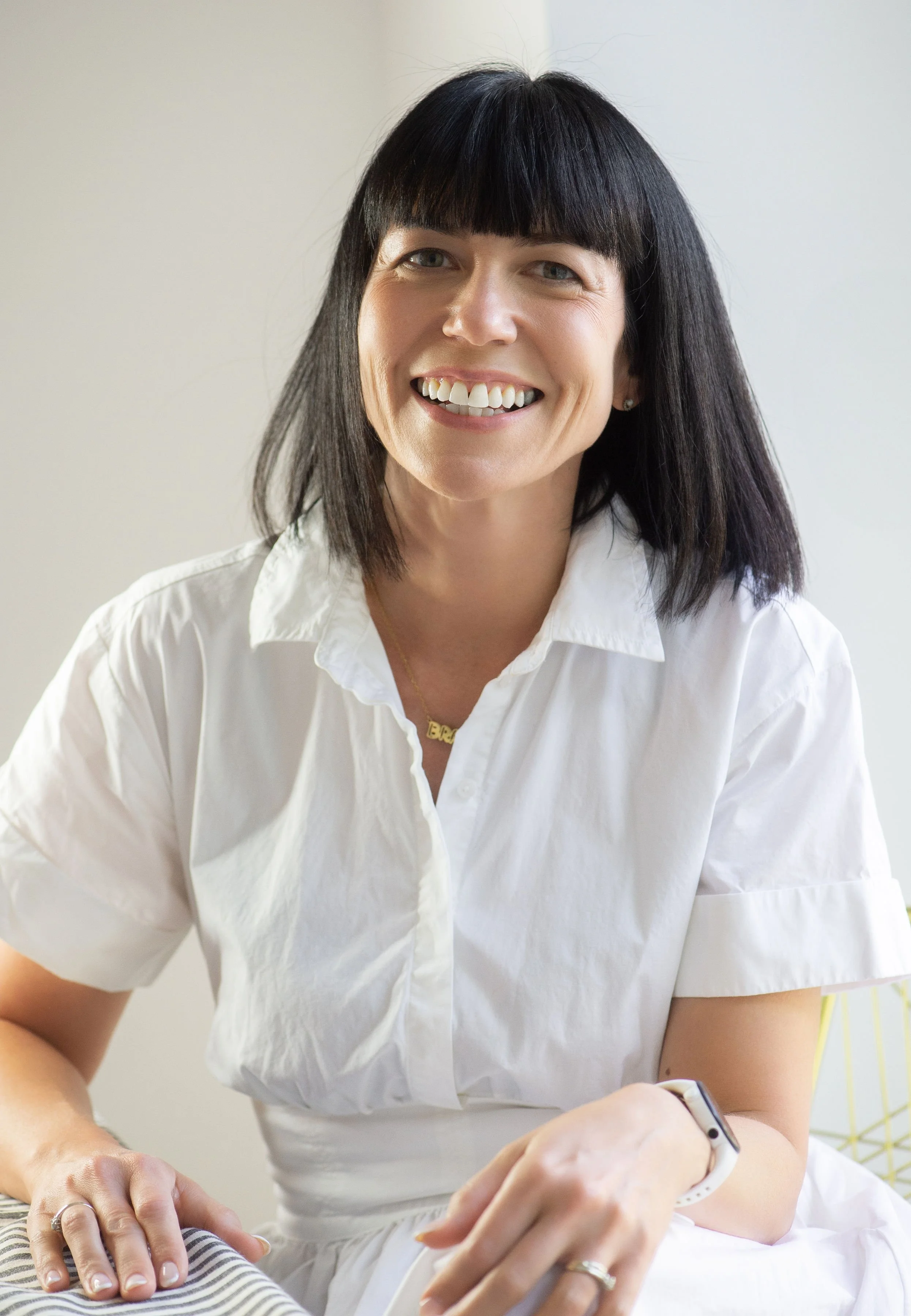 Close-up of a smiling woman with dark hair, wearing a white shirt, sitting at a table with her hands visible, one with a ring, and wearing a smartwatch on her wrist. The background is neutral and minimal.
