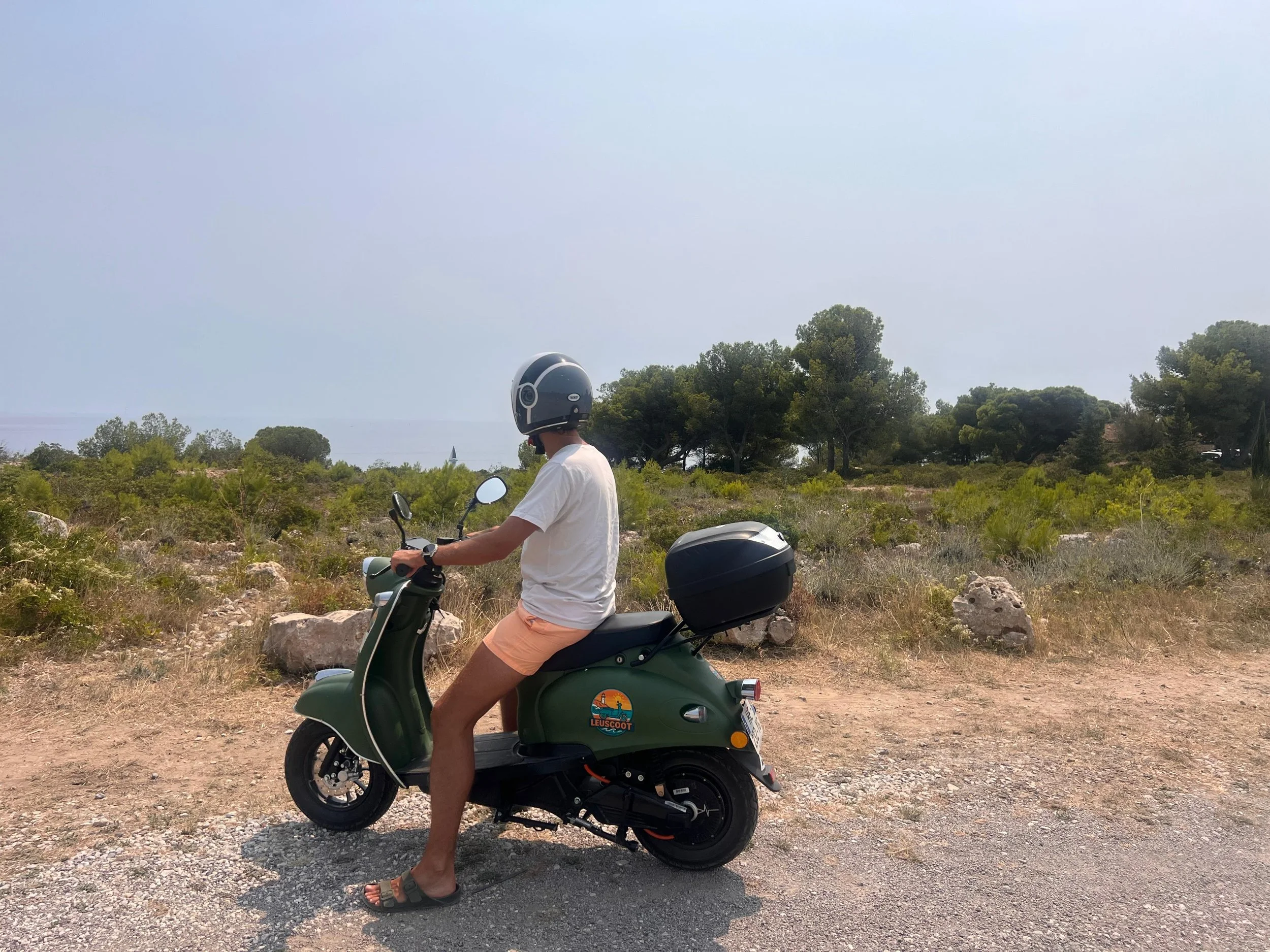 Person wearing a white T-shirt, peach shorts, sandals, and a helmet sitting on a green scooter with a back storage box, parked on a dirt path in a semi-arid landscape with shrubs, rocks, and trees under a cloudy sky.