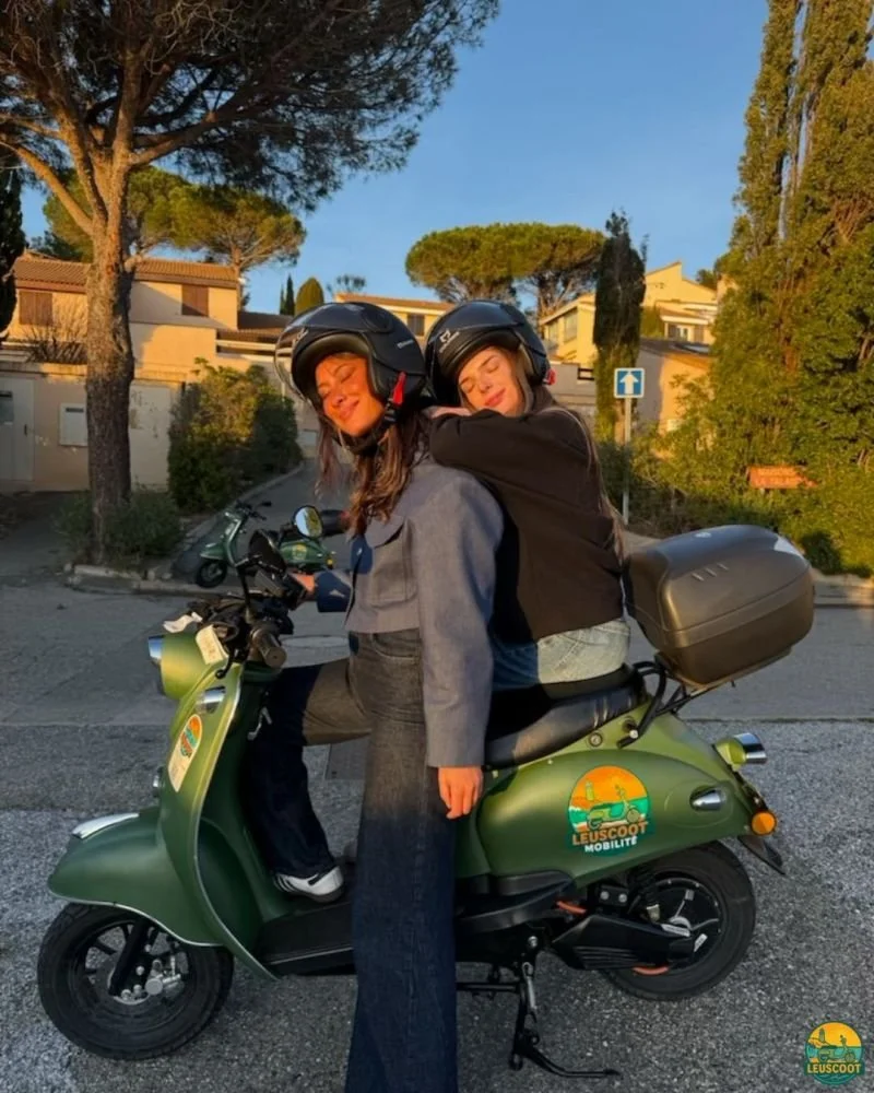 Two women wearing helmets sitting on a green scooter with a 'LEUSCOOT' logo, parked on a street with trees and houses in the background during sunset.
