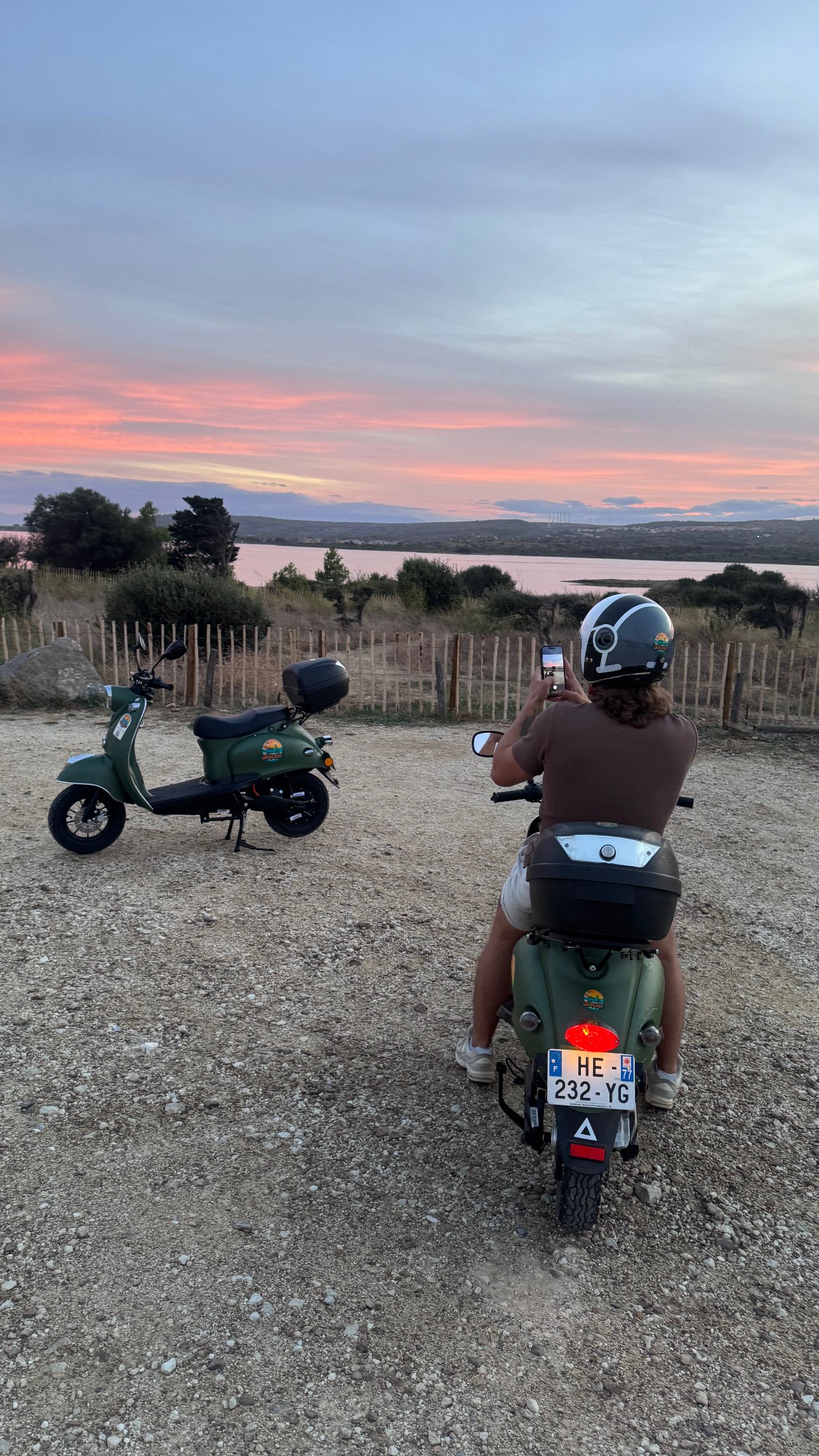 Person sitting on a green scooter taking a photo of the sunset over a lake, with a second scooter parked nearby and animals in the background.