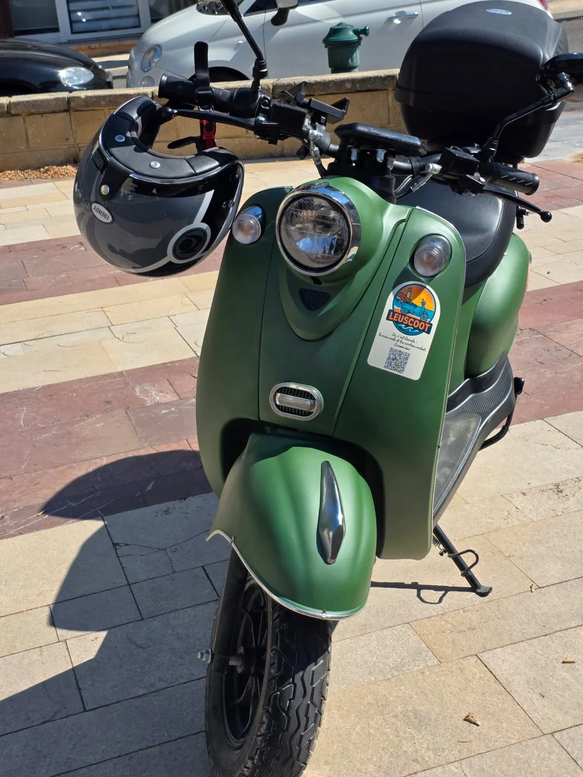 A green electric scooter with a black storage box on the back, parked on a sidewalk. It has a helmet hanging from the left handlebar and a sticker on the front with a logo that says 'LeusCoot' and features a cartoon of a person riding a scooter.