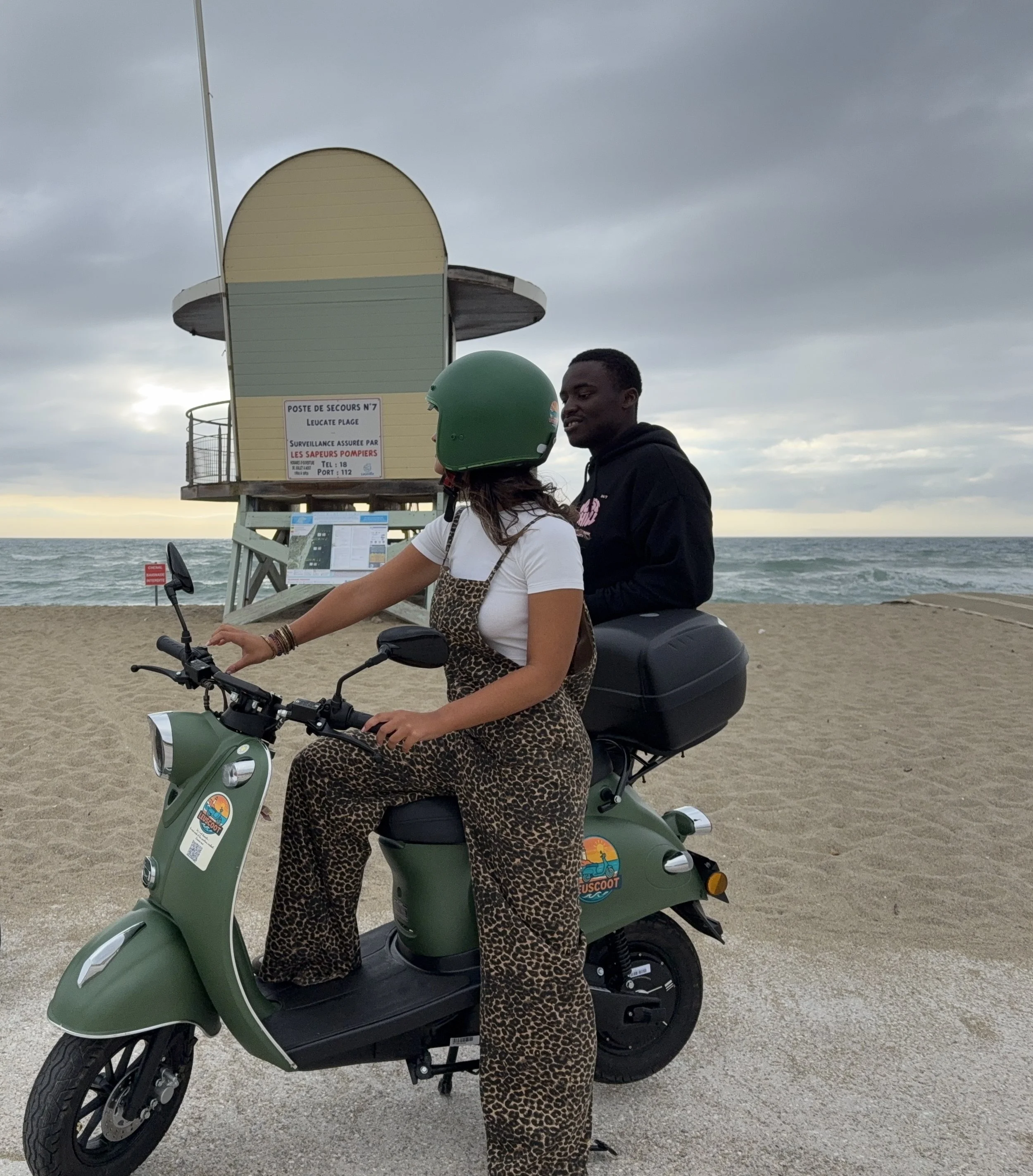 A woman wearing leopard print overalls and a green helmet riding a green scooter on a beach, with a man sitting behind her, in front of a lifeguard tower under cloudy skies.