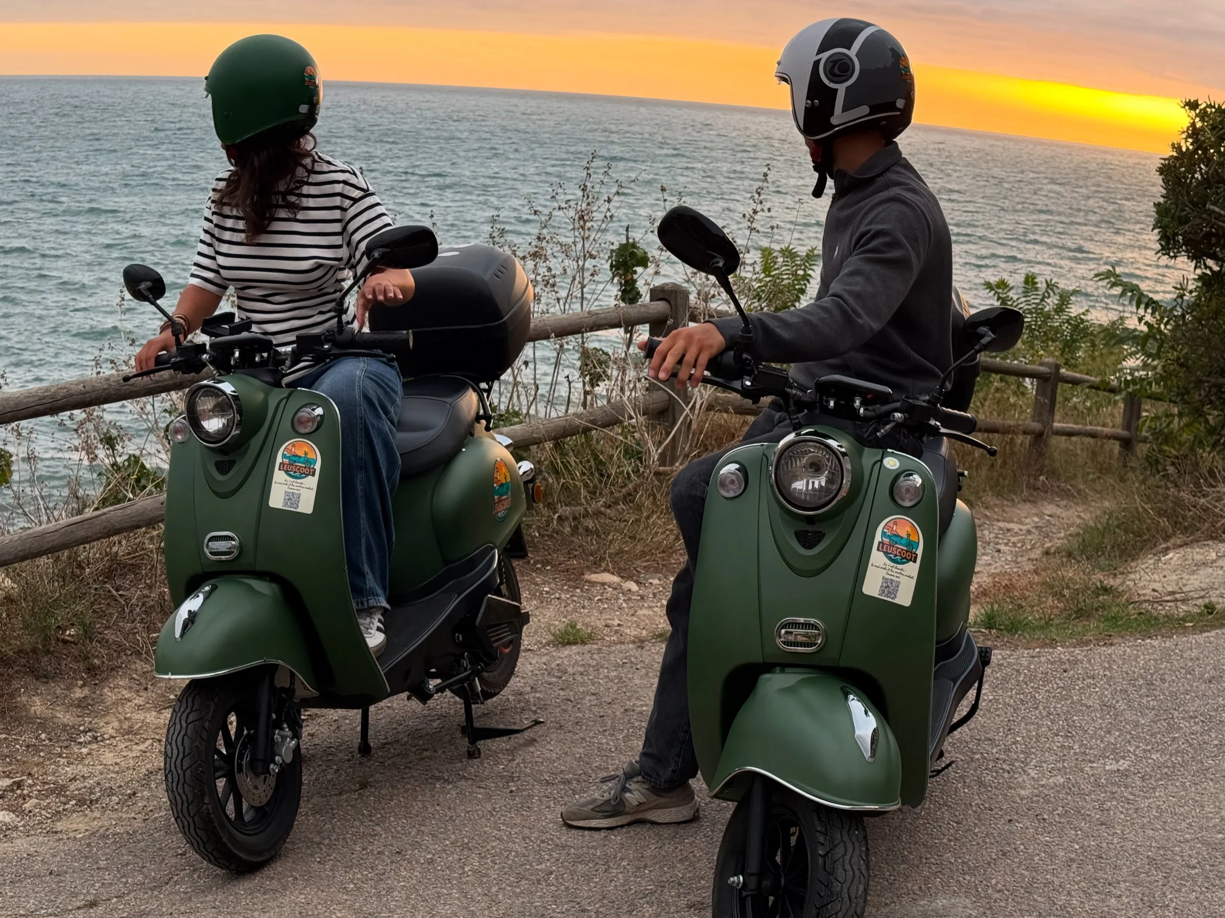 Two people wearing helmets riding green scooters along a coastal path at sunset, with a view of the ocean and sunset in the background.