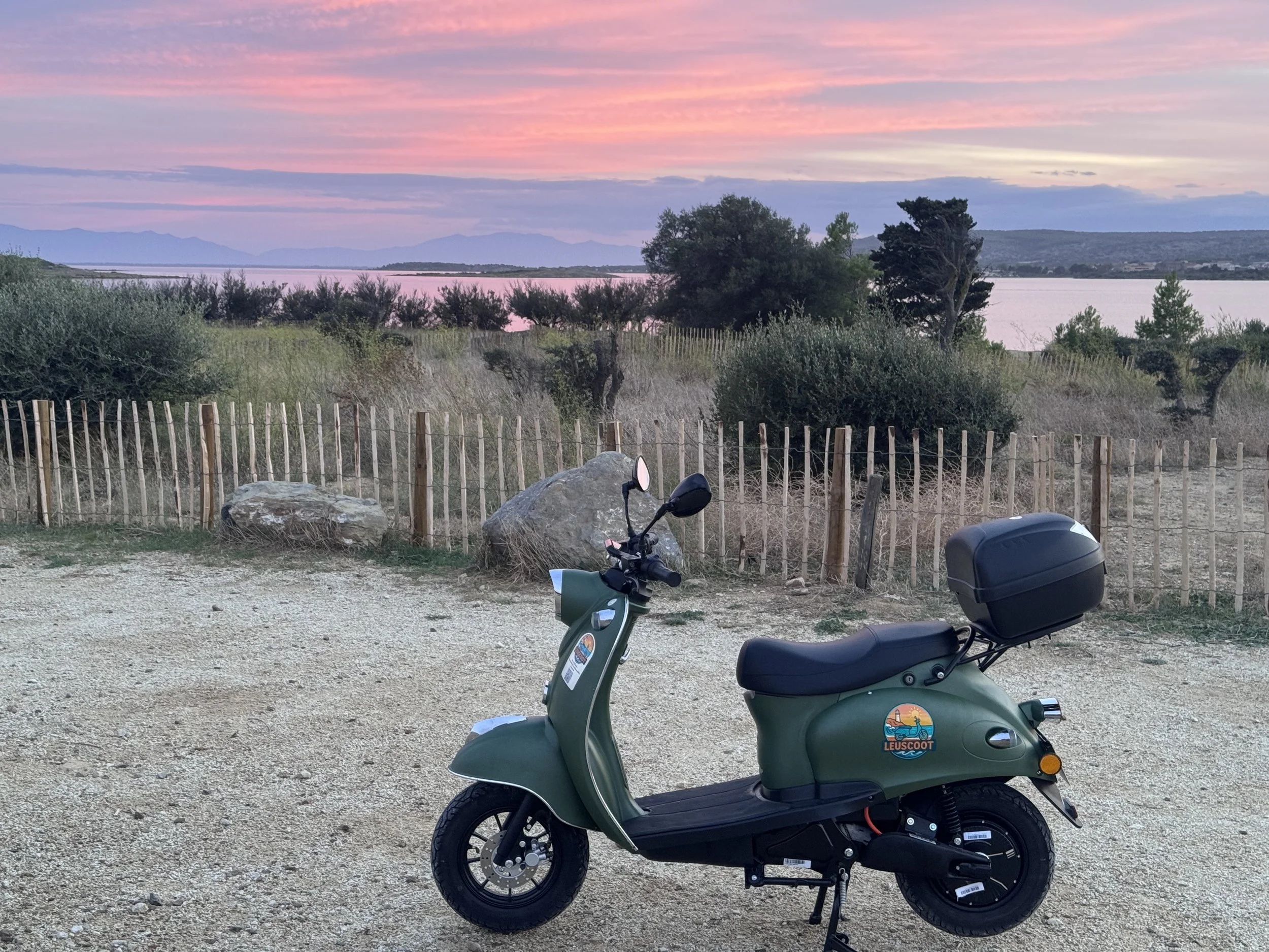 A green scooter parked on a gravel area near a wooden fence, overlooking a body of water at sunset with pink and purple sky, trees, shrubs, and mountains in the background.