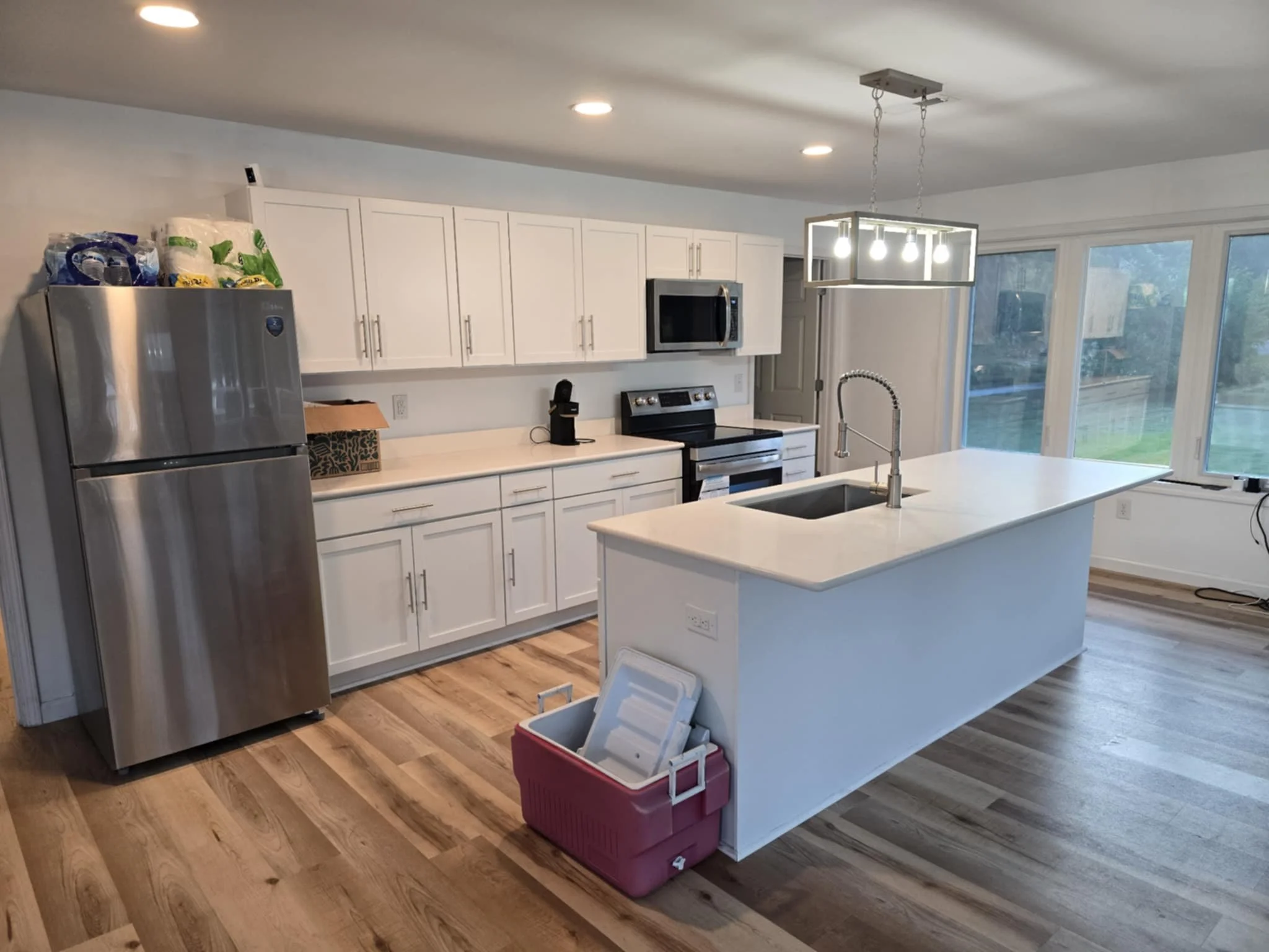 Modern kitchen with white cabinets, stainless steel refrigerator, black microwave and oven, island with sink, large windows, hardwood floors, and a hanging light fixture. We serve homeowners and businesses in Madison, Hanover, Scottsburg, Austin