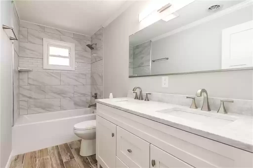 Bathroom featuring a bathtub with a gray marble tiled wall and a small window, a white vanity with a dual sink and silver fixtures, a large mirror, and wood-look tile flooring. We serve homeowners and businesses in Madison, Hanover, Scottsburg