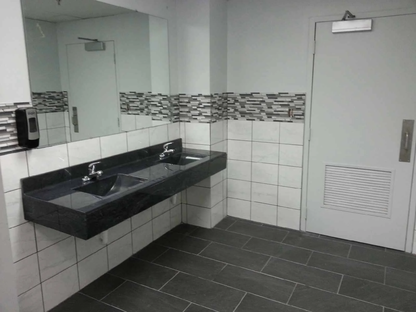 Public restroom with a double sink, large mirror, soap dispenser, and tiled walls in shades of gray and white. We serve homeowners and businesses in Madison, Hanover, Scottsburg, Austin, Milton, KY, Carrollton, KY and surrounding communities 