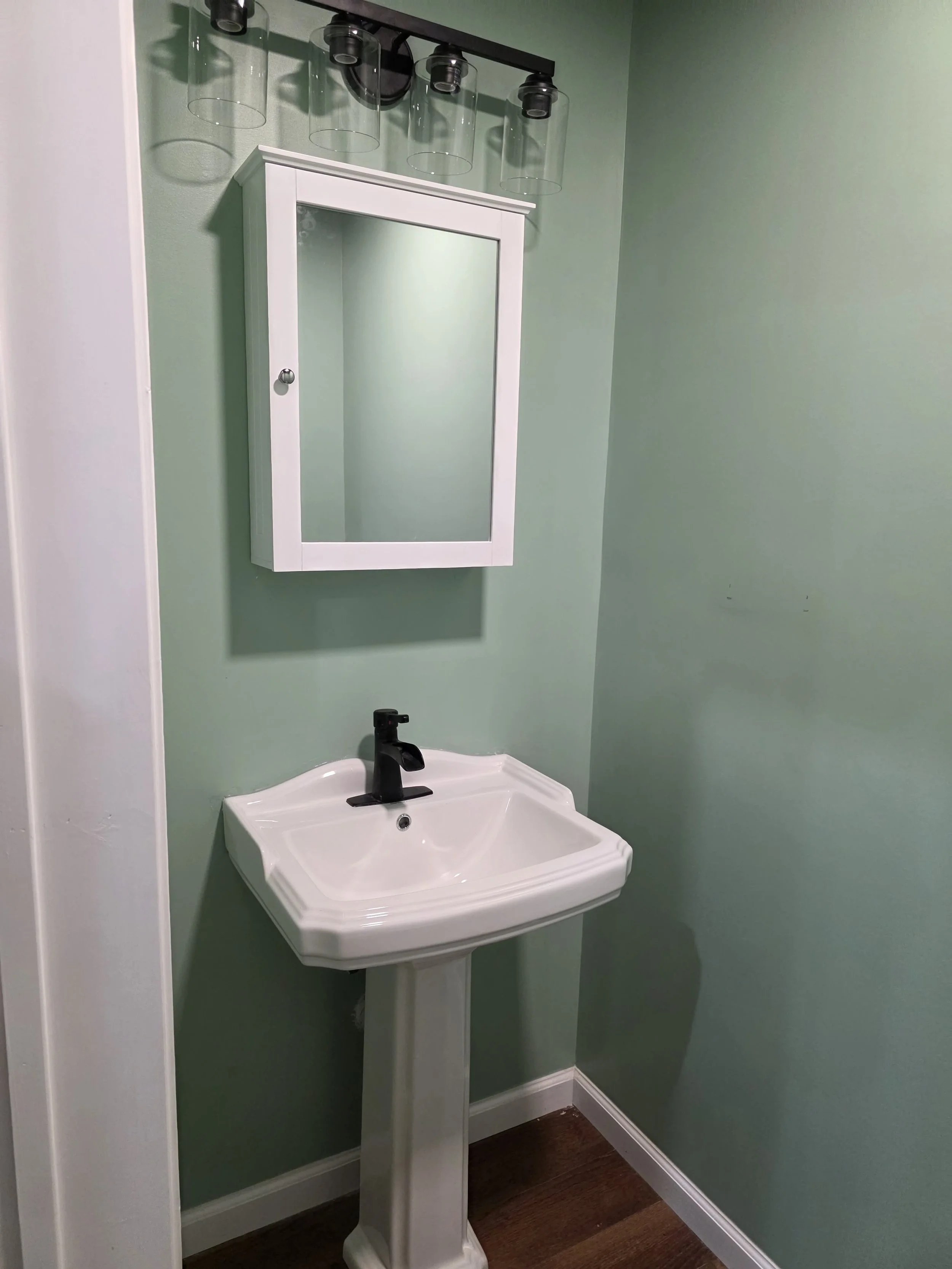 A small bathroom with green walls, a white pedestal sink with a black faucet, a white medicine cabinet with a mirror, and a light fixture with four clear glass shades above the mirror. We serve homeowners and businesses in Madison, Hanover