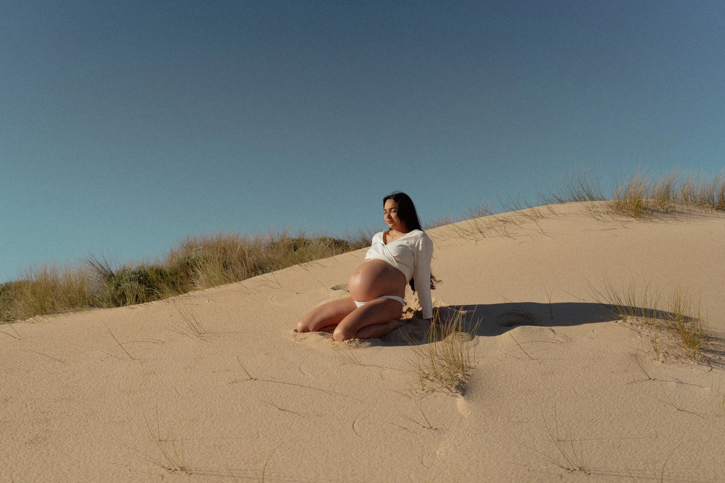 A relaxed maternity shoot with a pregnant woman enjoying a quiet, reflective moment sitting on sand dunes with patches of grass under a clear blue sky.