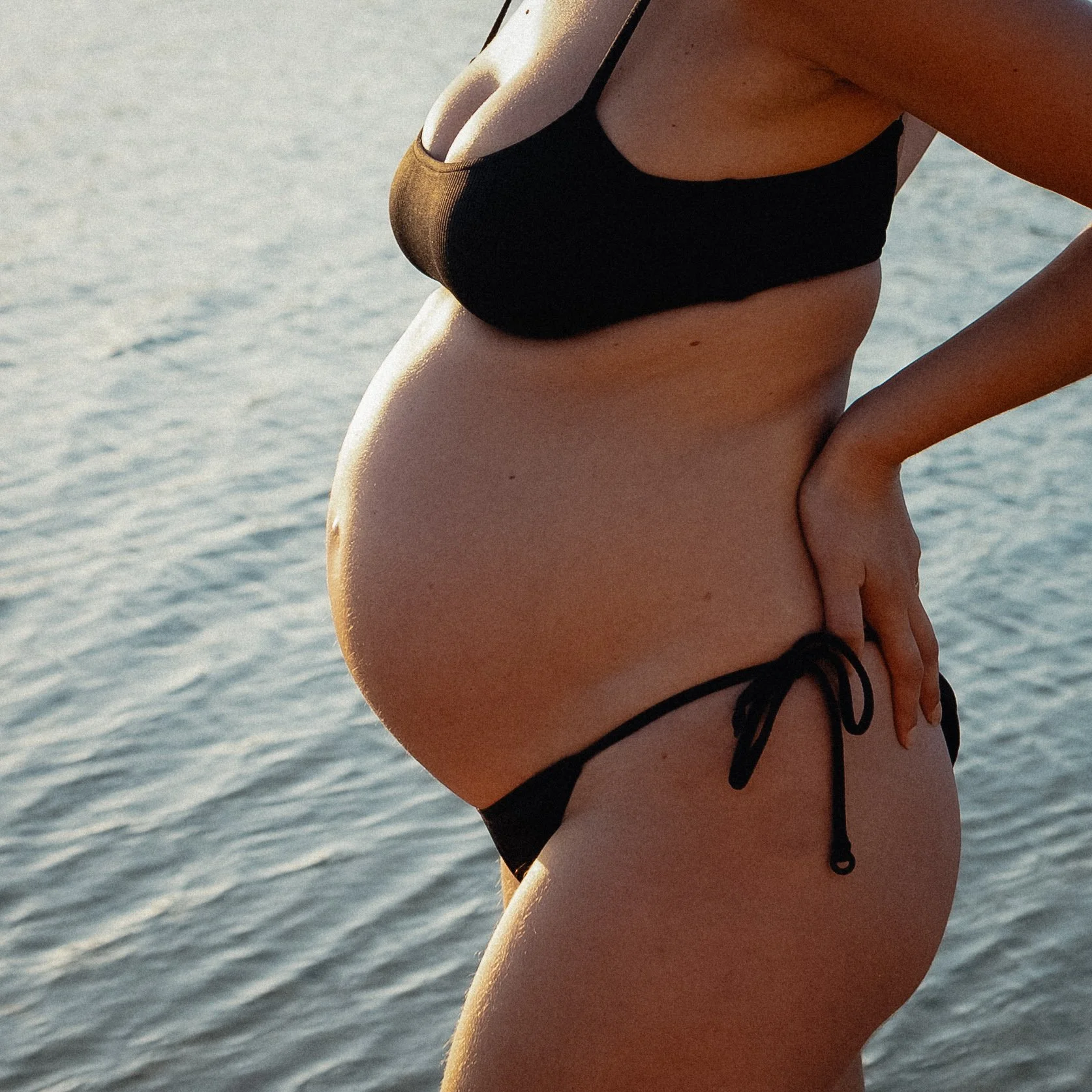 Golden sunlight illuminates an expectant mother in a black bikini beside the river at the Margaret River Mouth, photographed during a relaxed Margaret River maternity photography session.