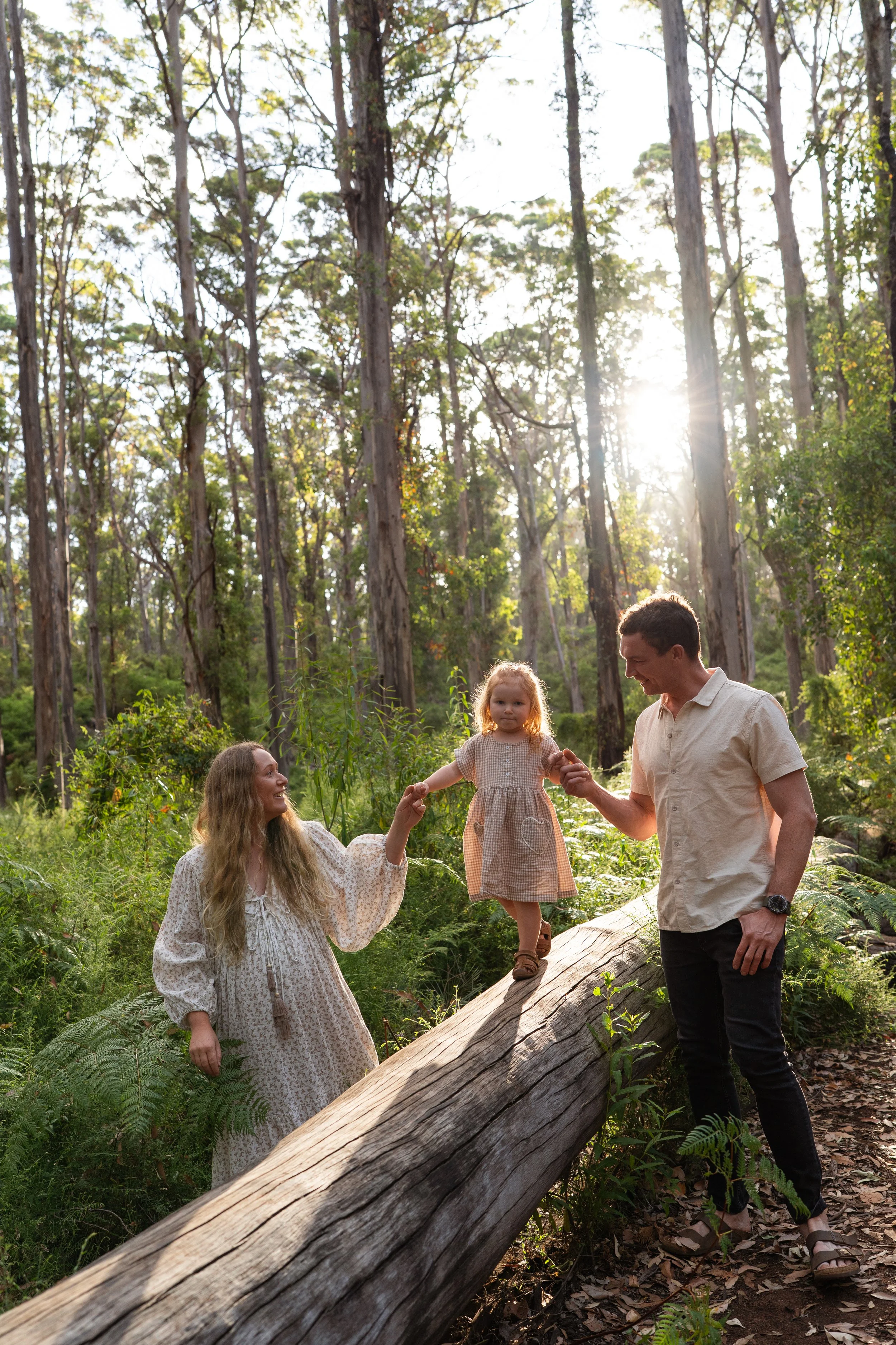 A family of three enjoying time in the Boranup forest in Margaret River during a relaxed family photoshoot. The mother is on the left, the father on the right, and their young daughter is standing on a fallen log between them. The family members are 