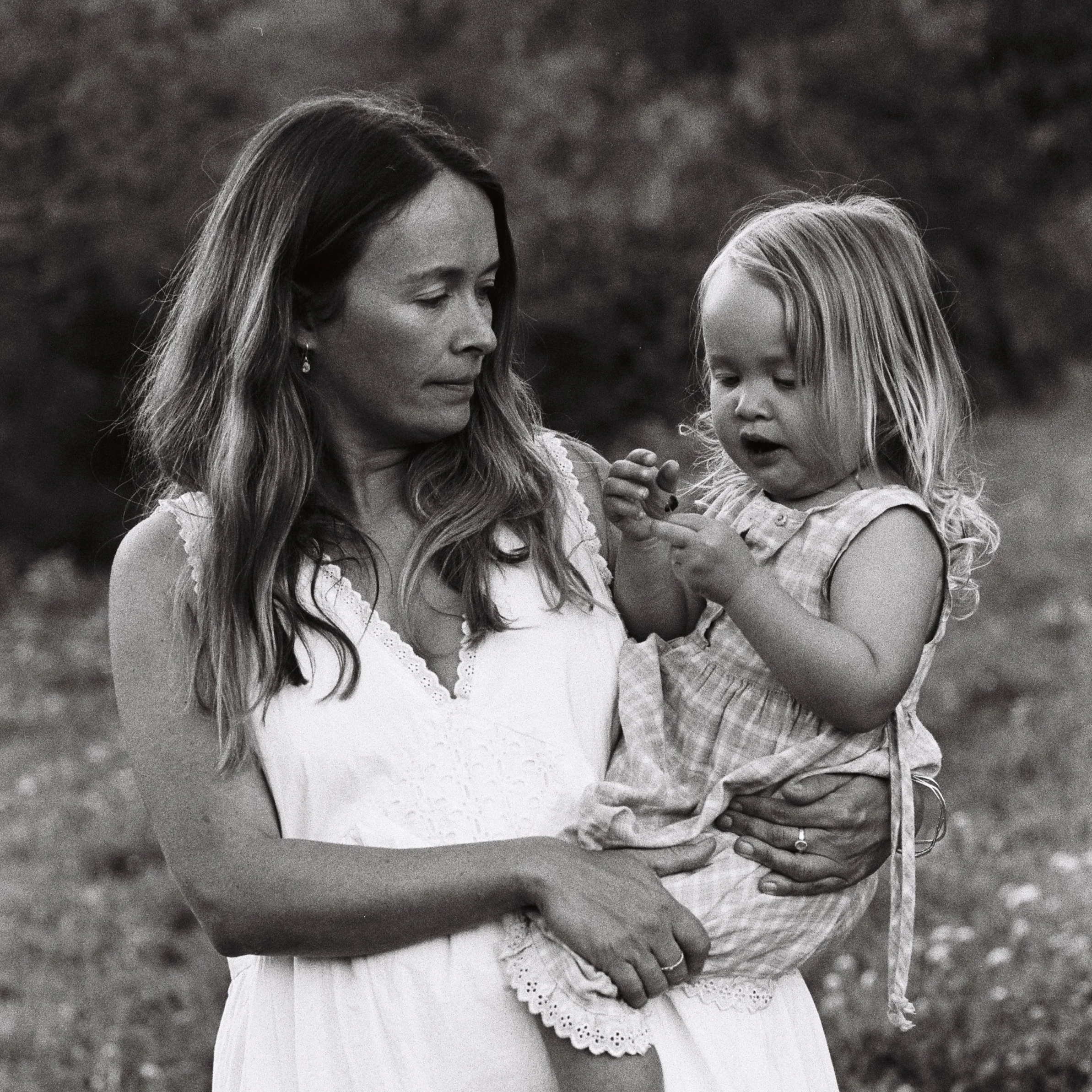 Black and white photo of a woman and a young girl in a grassy outdoor setting. The woman has long, wavy hair and is wearing earrings and a dress with lace details. The girl has long hair and is looking down while holding the woman's hand. The scene appears to capture a tender moment between them.