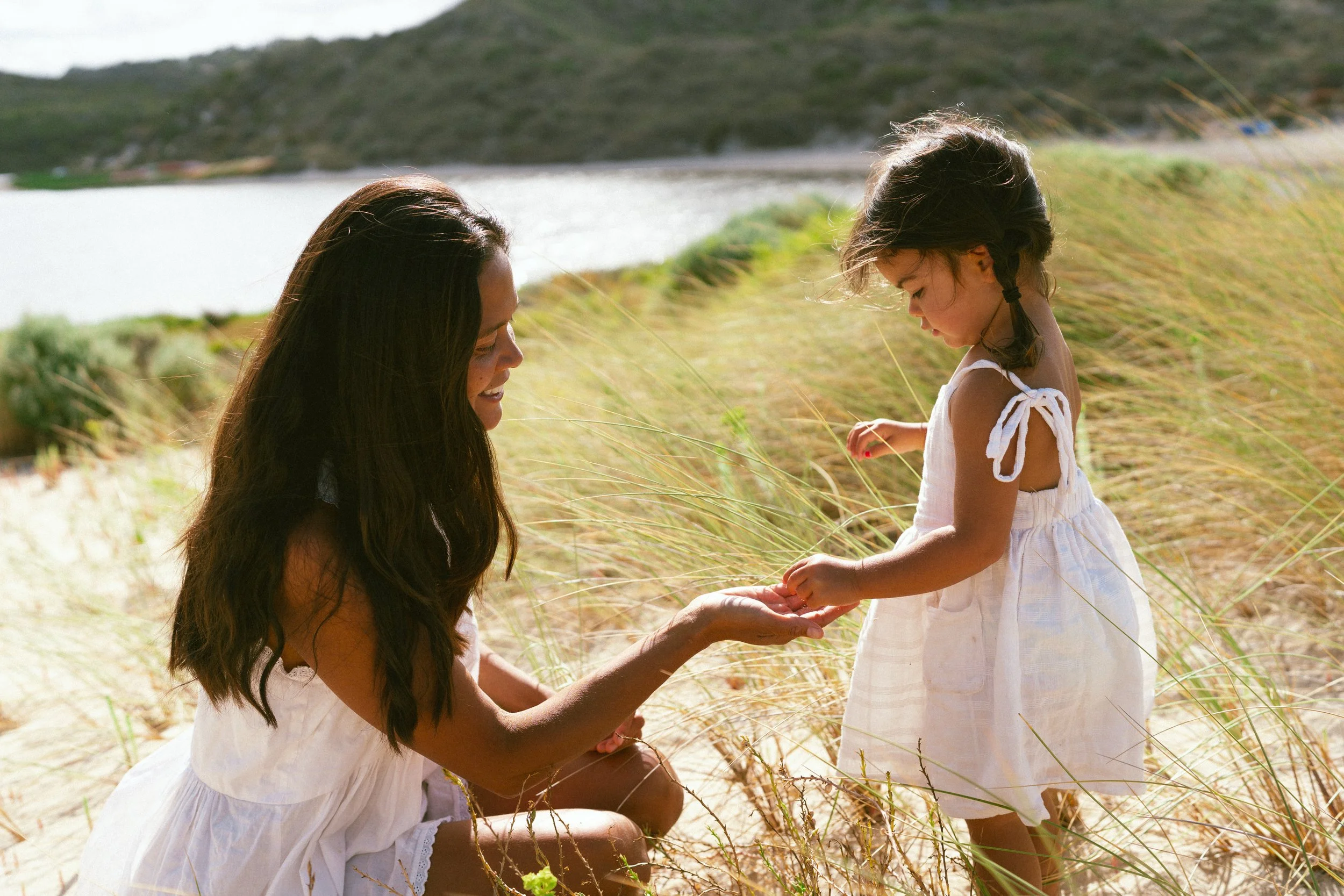 A mother and daughter during a motherhood family photoshoot in white dresses on a beach with tall grass, holding hands and looking at each other in a relaxed and connected moment. The background features the Margaret River Rivermouth.