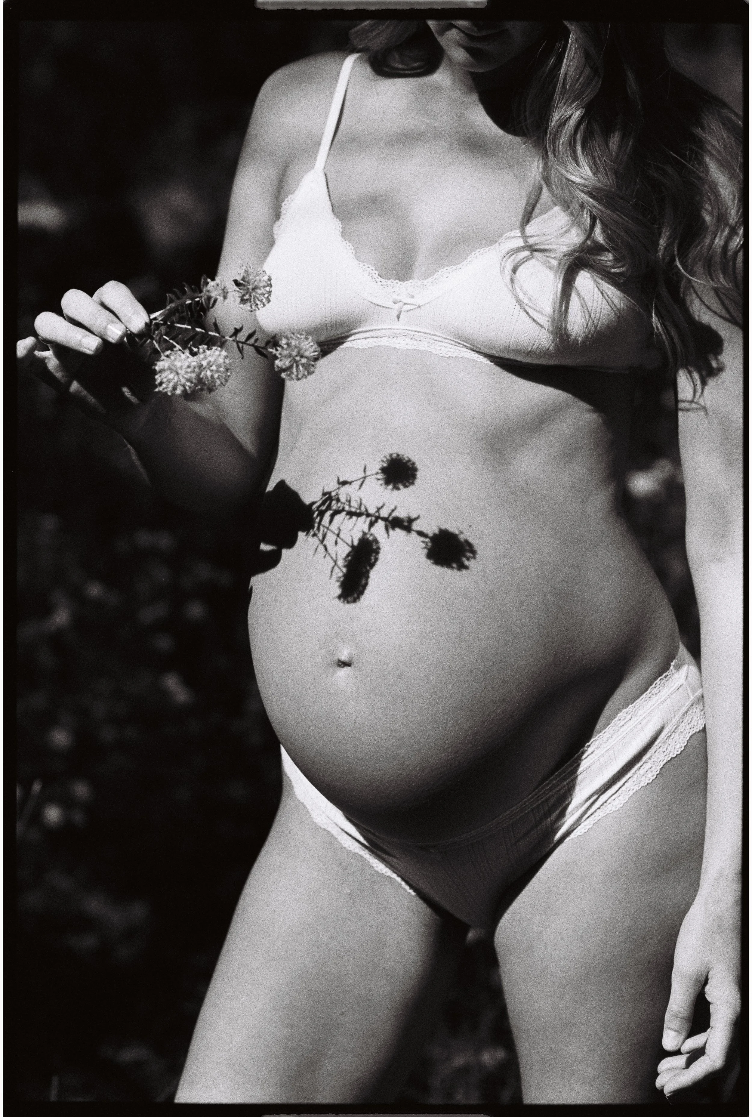 Black and white 35mm film maternity photo of a pregnant woman in lingerie holding flowers, with the flowers casting a shadow on her pregnant belly.