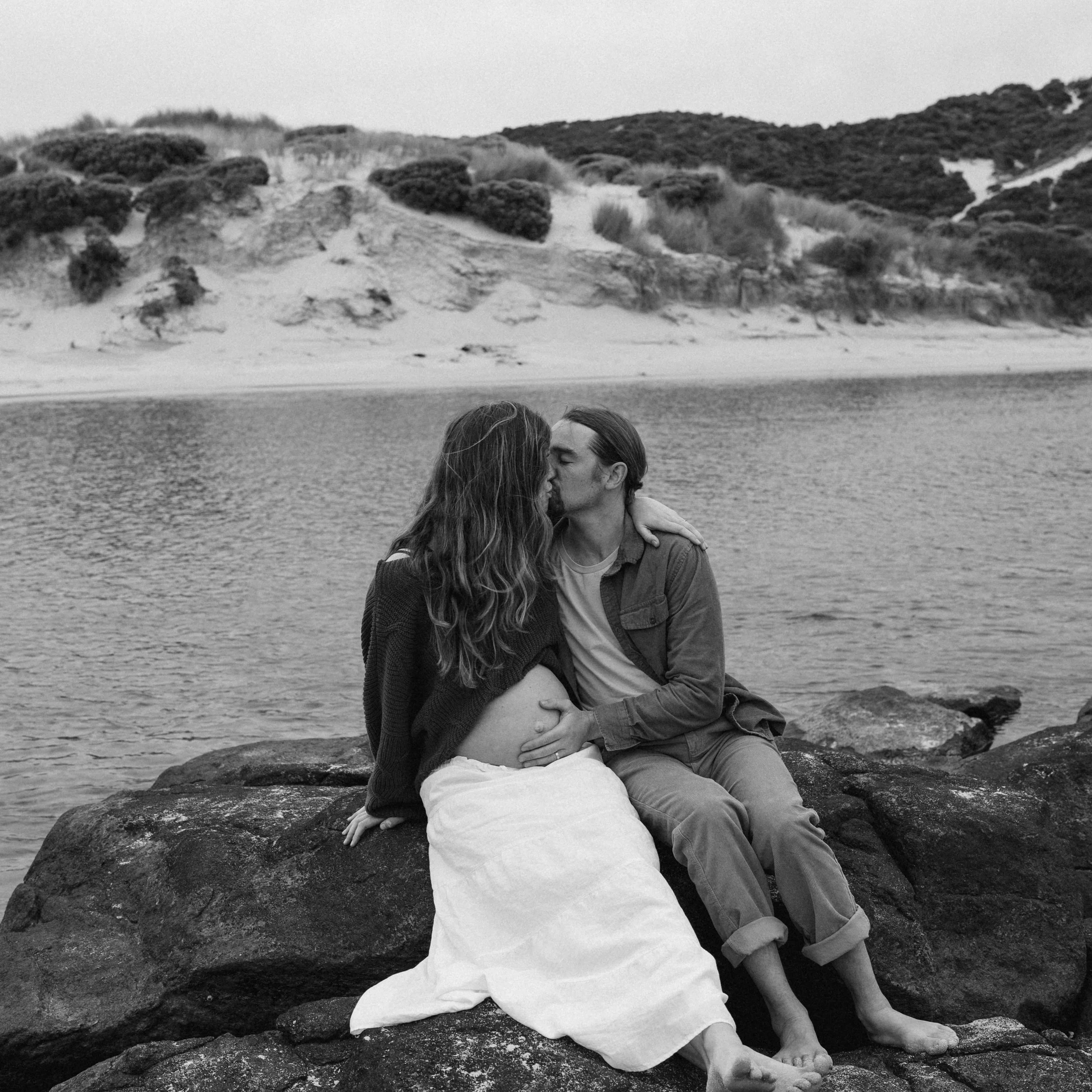 A black-and-white photo of a couple at a couples maternity photoshoot sitting on rocks by the water by the Margaret River Rivermouth, sharing an intimate kiss; the woman is pregnant and holding her belly, while the man holds her close at a beach with