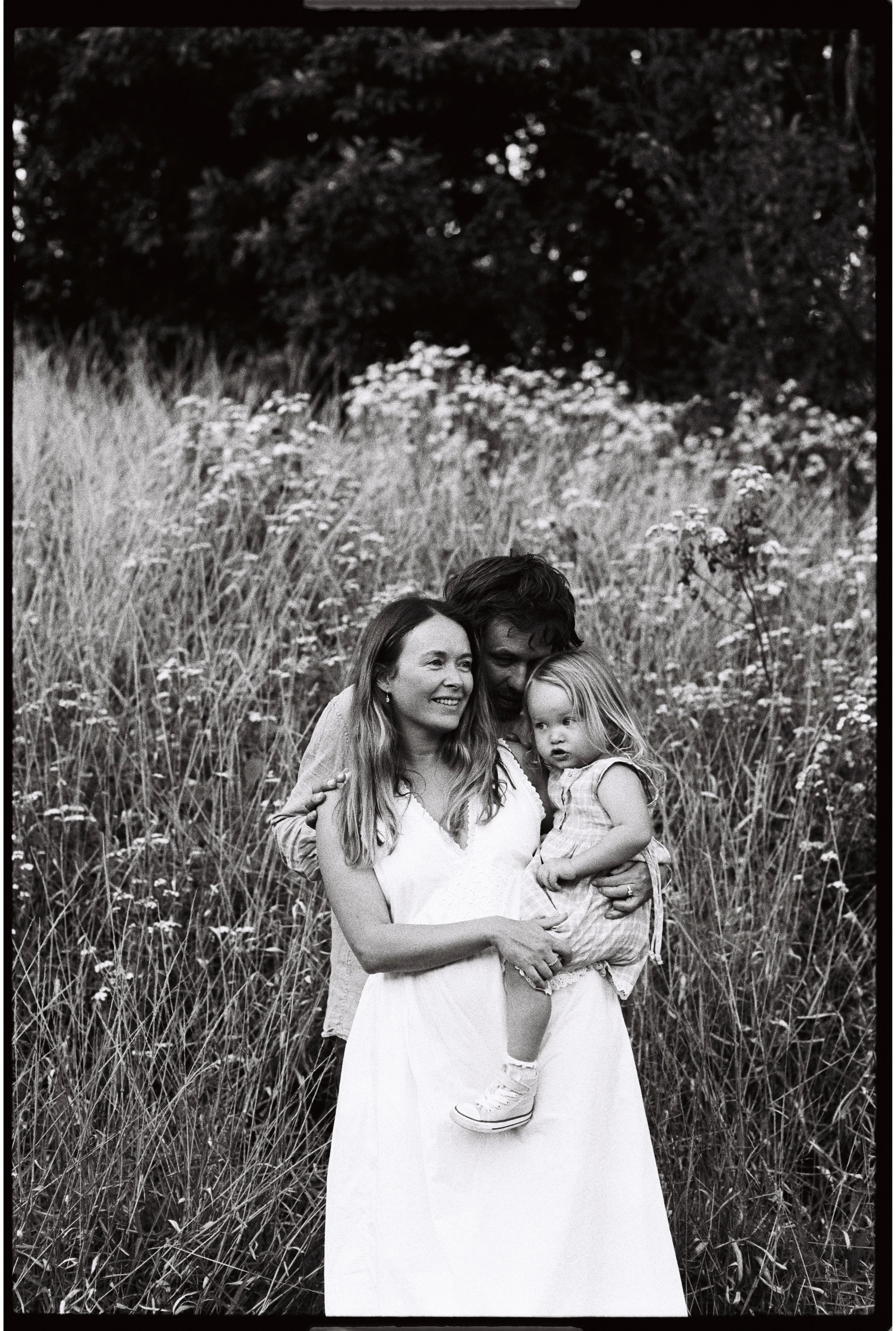 A black and white film photo on 35mm of a mother holding her daughter with the father standing behind them. All cuddled together with tall grass and flowers behind them