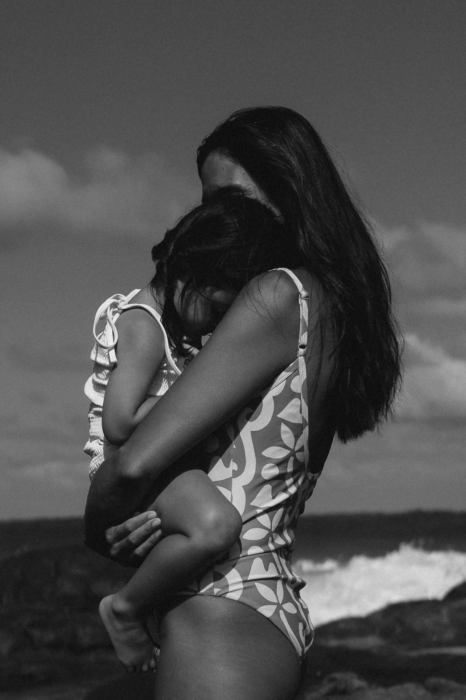 A woman holding a child near the ocean on a cloudy day, both with dark hair, in black and white.
