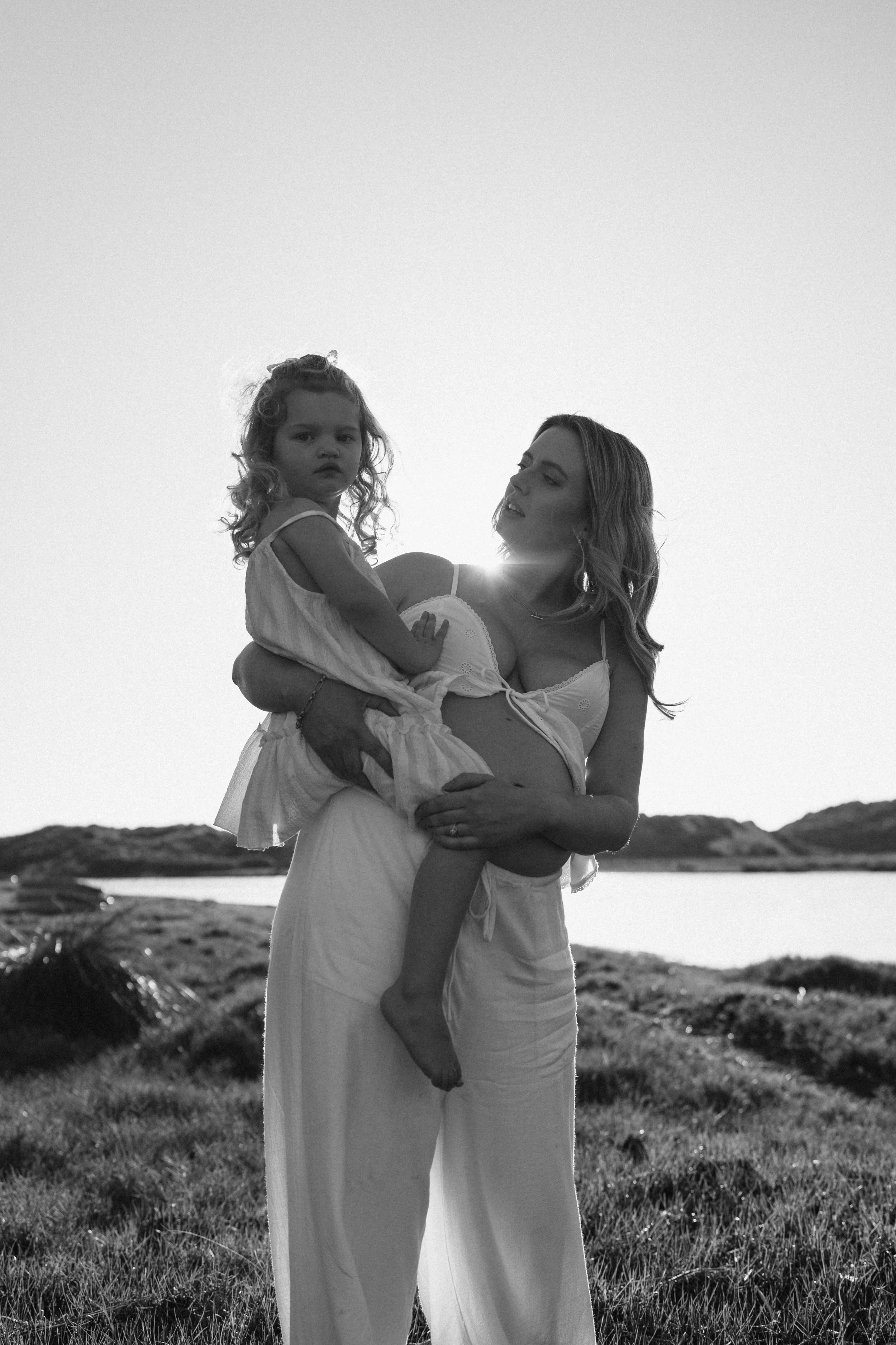 A mother and daughter sharing a playful, connected experience outdoors during a motherhood maternity shoot at the Margaret River Rivermouth. The scene is backlit by the sun, creating a silhouette effect in black and white.
