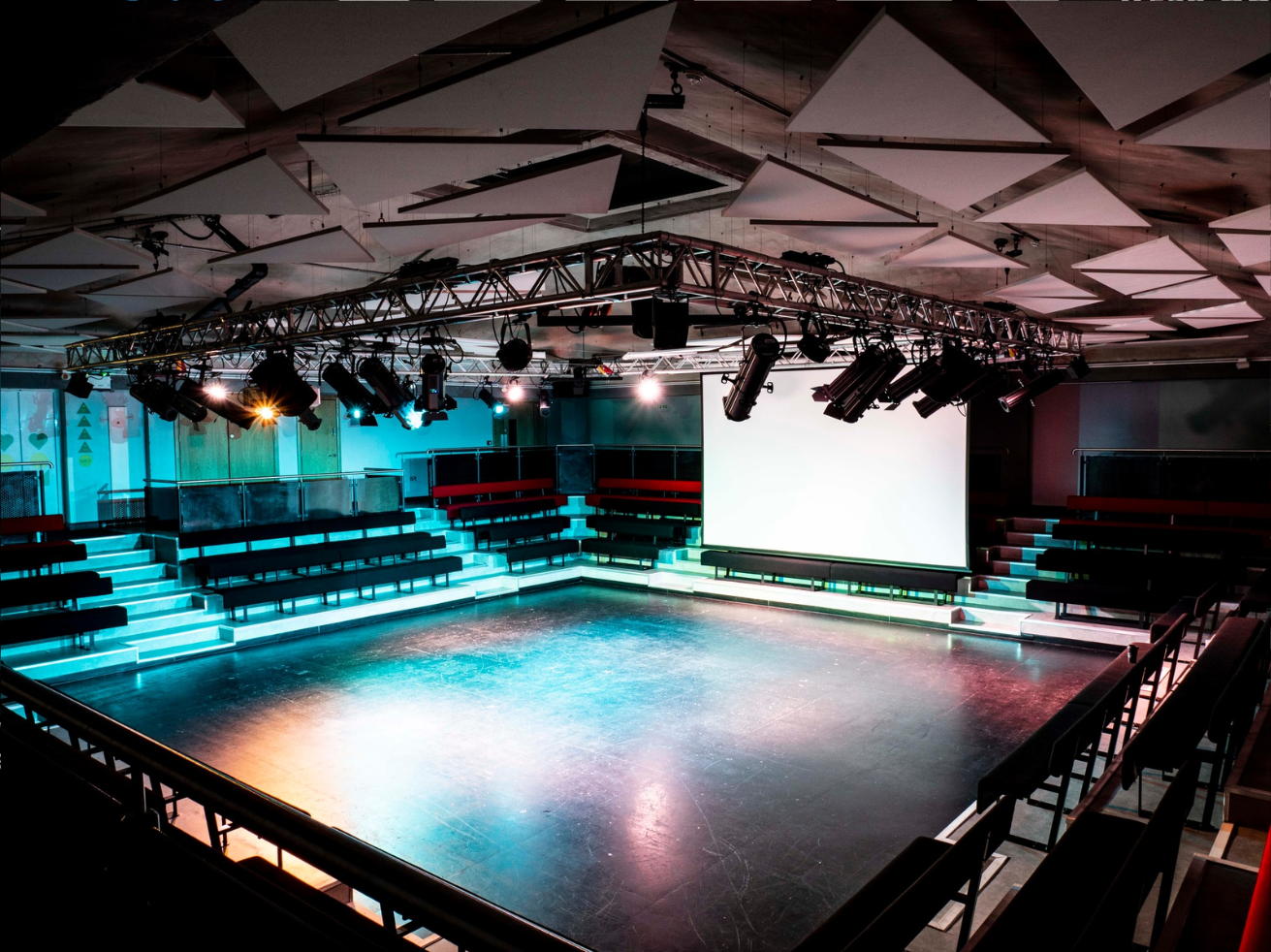 An empty theater or performance space with tiered seating, stage lighting, a large projection screen, and a dark polished floor.