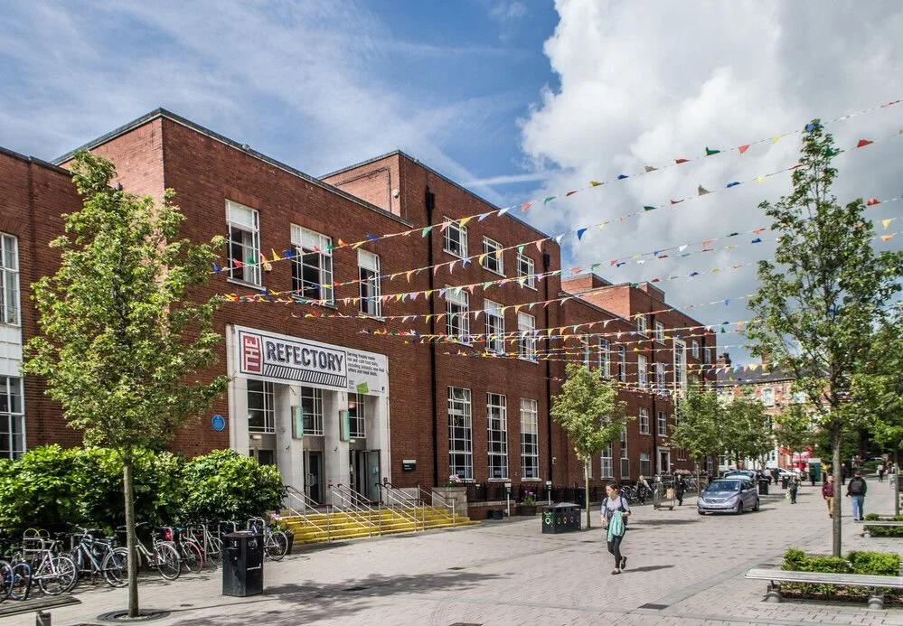 A brick building with a sign that says "The Refectory," decorated with colorful pennant flags, situated along a pedestrian-friendly street with trees, bicycles, and people walking.