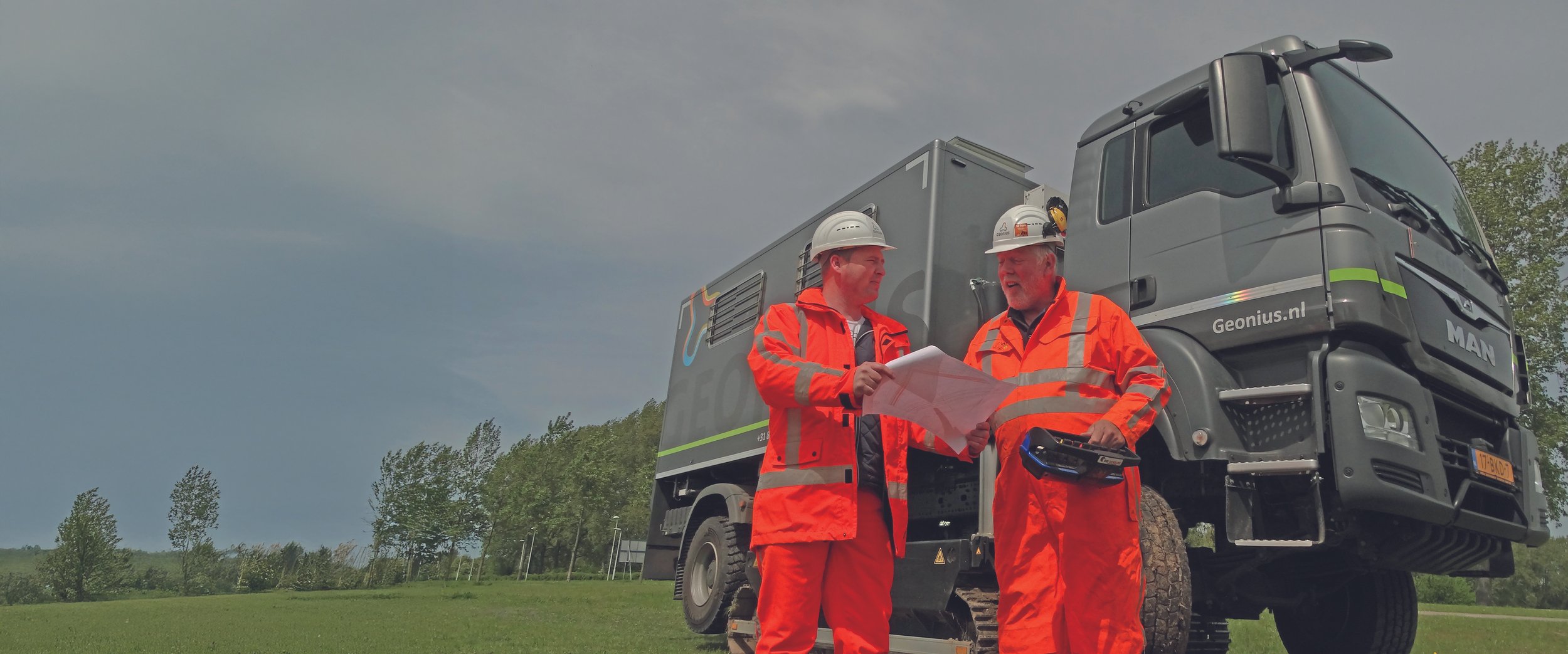 Twee mannen in oranje veiligheidskleding en helmen die op een veld staan, bespreken plannen naast een groot technisch voertuig.