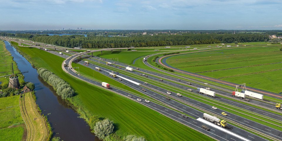 Luchtfoto van een snelweg met meerdere rijbanen en verkeer, omringd door groene weilanden en een rivier aan de linkerkant.