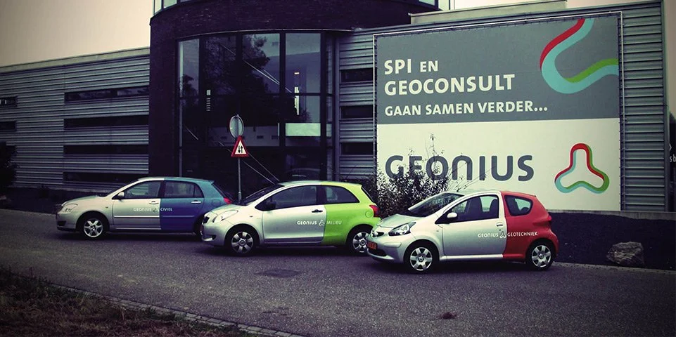 Drie auto's geparkeerd naast een gebouw met een groot reclamebord dat schrijft: "SPI EN GEOCONSULT Gaan Samen Verder... GEONIUS."