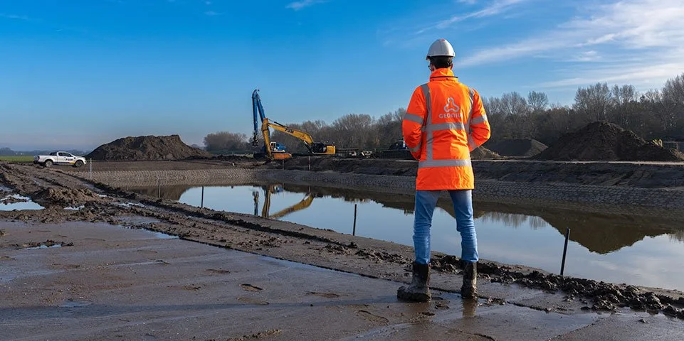 Een bouwvakker in een oranje veiligheidsvest en helm staat op een bouwplaats bij een waterpartij, terwijl een graafmachine op de achtergrond het terrein bewerkt.