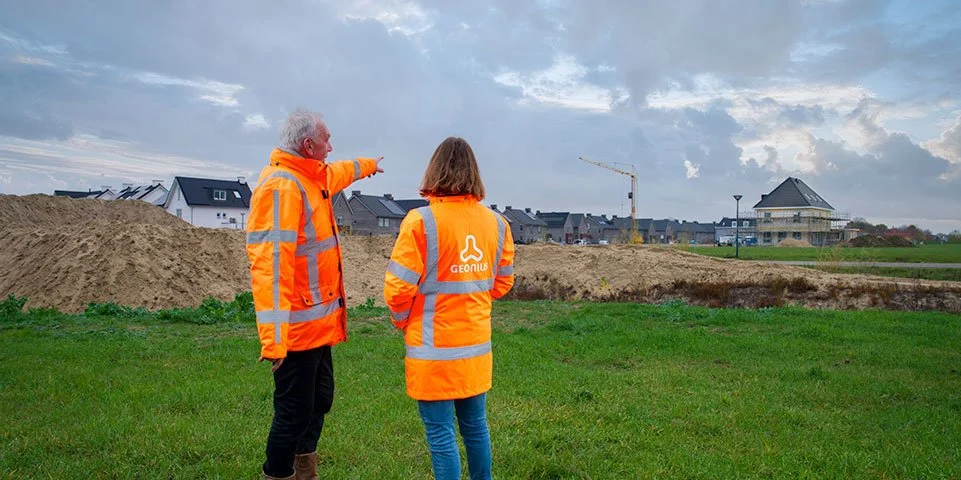 Twee mensen in oranje veiligheidsjassen staan op een grasveld bij een bouwplaats, waarbij een man wijst naar de bouwactiviteiten in de verte.