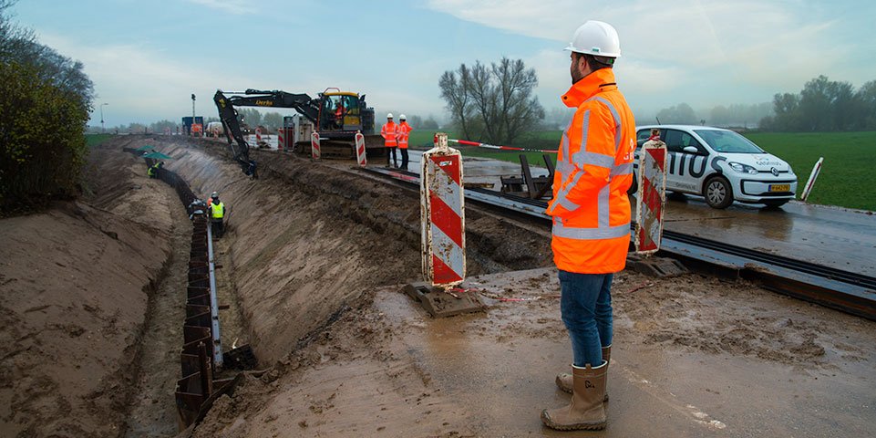 Arbeiders en een machine aan een weg- of spoorwegproject tijdens regenachtig weer, met bouwhekken en een auto op de achtergrond.