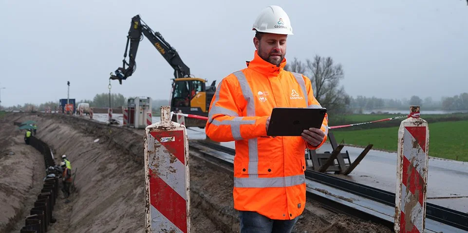 Een bouwvakker in een oranje jas en witte helm die een tablet vasthoudt op een bouwplaats met graafwerkzaamheden en machines.