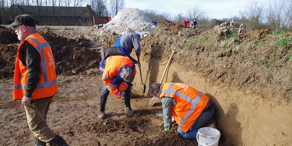 Mensen werken op een bouwplaats, graven in de grond met behulp van hamers en scheppen, sommigen dragen oranje veiligheidshesjes.
