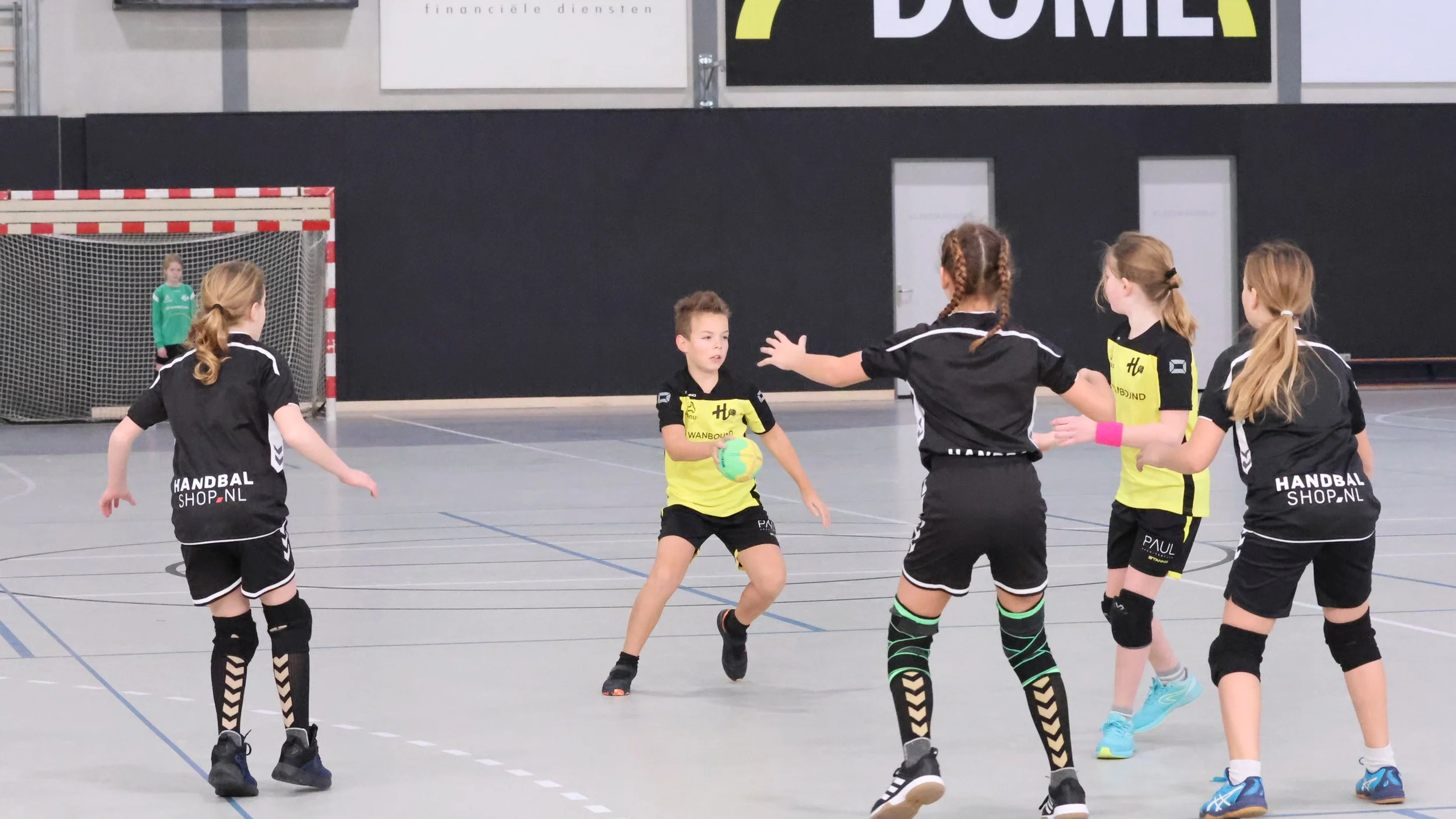 Jeugdige handbalspelers in een sporthal tijdens een wedstrijd, met de ene speler met de bal en de andere spelers die proberen te blokkeren. Achter hen een keepster bij het doel.