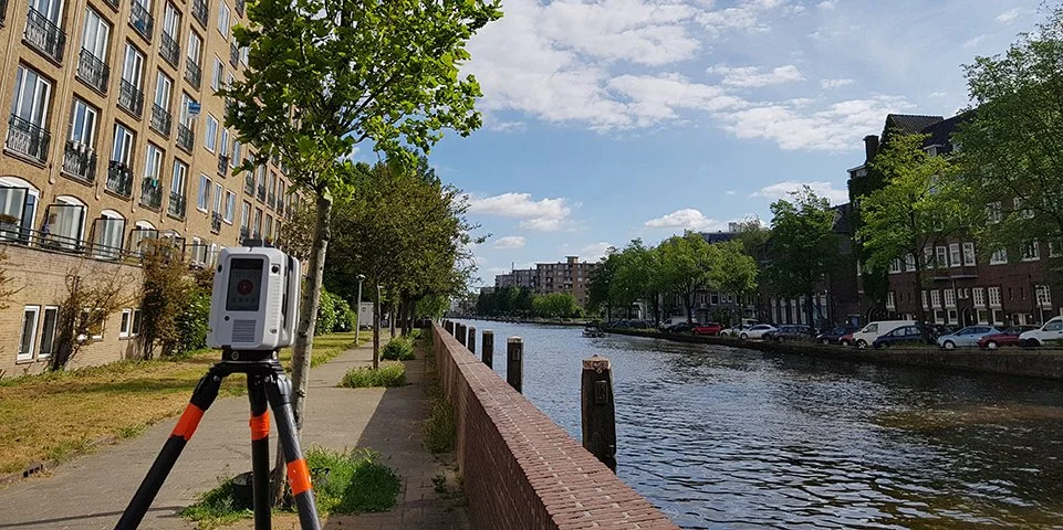 Stadsgezicht met water, wandelpad, bomen en gebouwen, een meetinstrument op statief langs de waterkant.