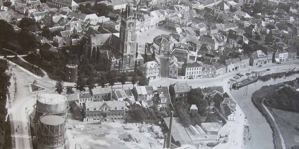 Luchtfoto van een historisch stadje met een kerk, woonhuizen, een kanaal met boten, en industriële structuren.