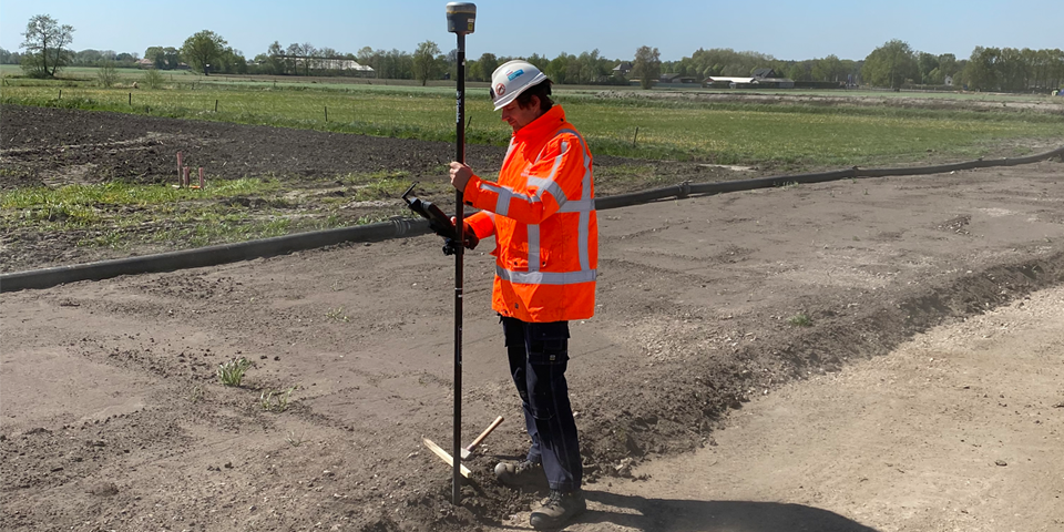 Een man in een oranje veiligheidsjas en helm gebruikt een GNSS-ontvanger op een landbouwveld.