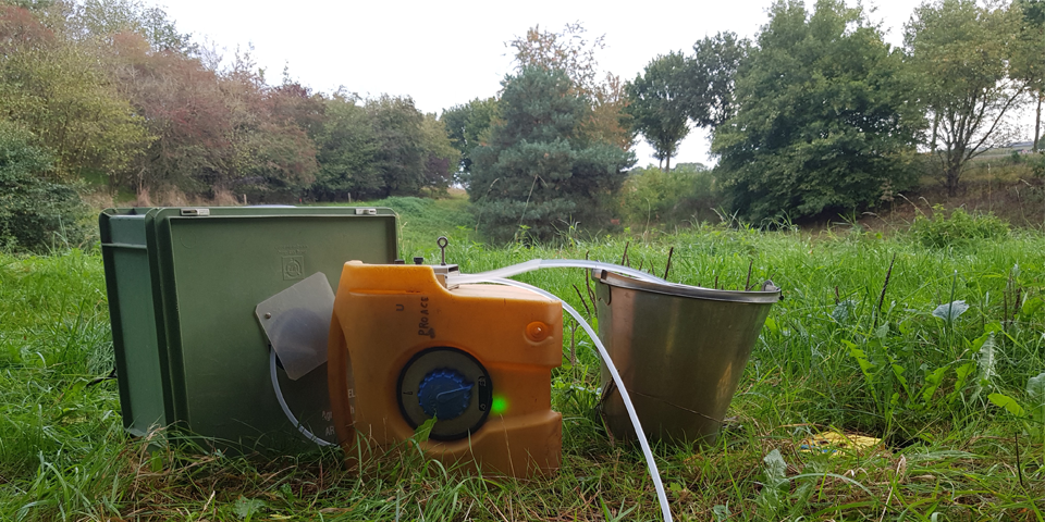 Een watermonsternemingsset met een milieuvriendelijke meetdoos, een waterbrug, en een metalen emmer, geplaatst op een grasveld met bomen op de achtergrond.