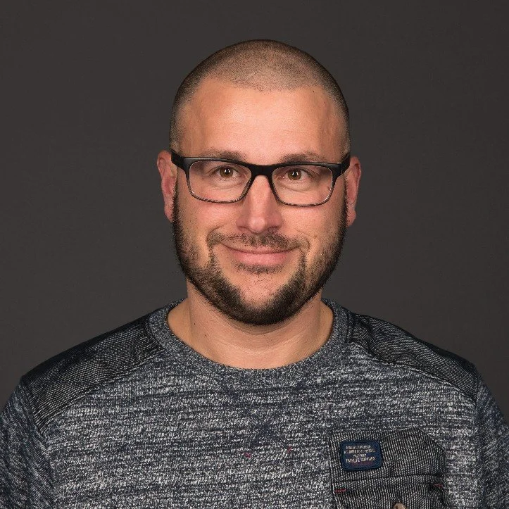 Portret van een man met een baard, bril en korte haar op een grijze achtergrond, lachend naar de camera.