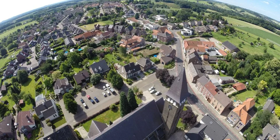 Luchtfoto van een woonwijk met diverse huizen, groene tuinen, een parkeerplaats, en omliggende landbouwvelden.