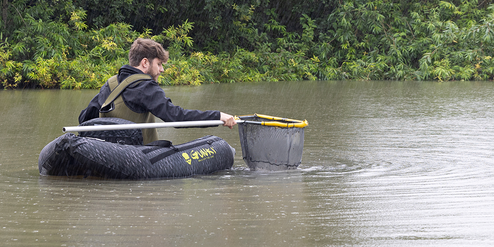 Man in een opblaasbare boot met een visnet in de rivier, omringd door groene planten.