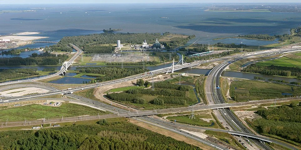 Luchtfoto van snelwegknooppunt met meerdere bruggen, wegen en groenvoorzieningen, watersysteem en industriegebied op de achtergrond.