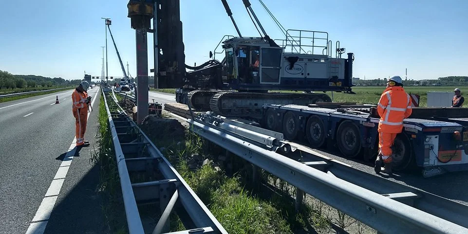 Terreinwerkers in heldere oranje kleding bij een wegwerkproject met een grote kraan en transportvoertuig op een snelweg in de branding van de zon.