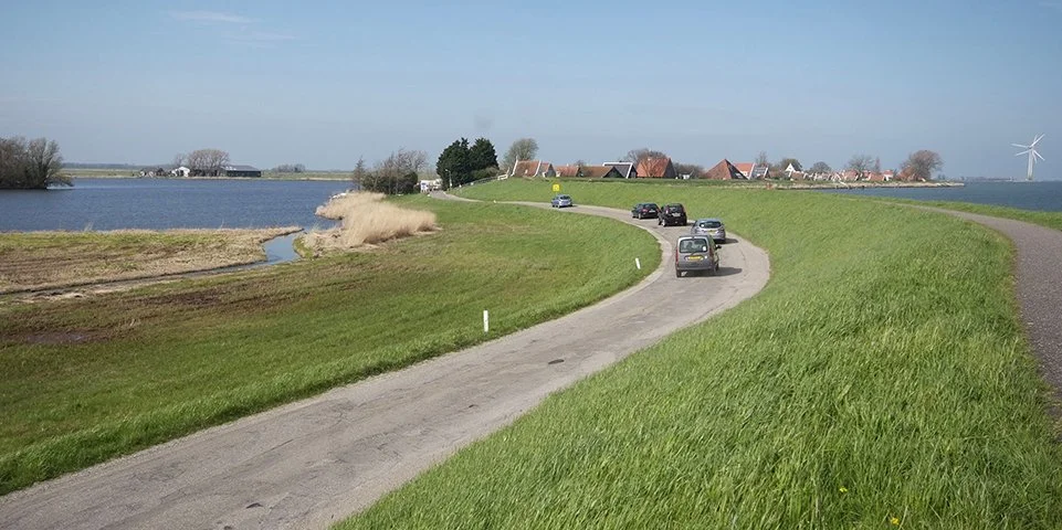 Zicht op een landelijke weg langs een meer met groene weiden en huizen op de achtergrond, windmolen op de horizon.