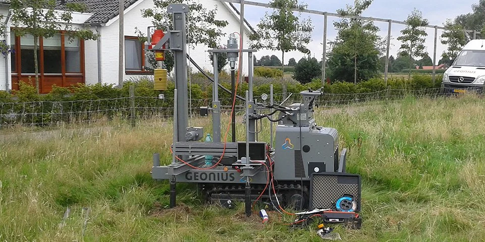 Een grijze geologische sonde met naam 'GEONUS' op een grasveld, met meetapparatuur en kabels eromheen.