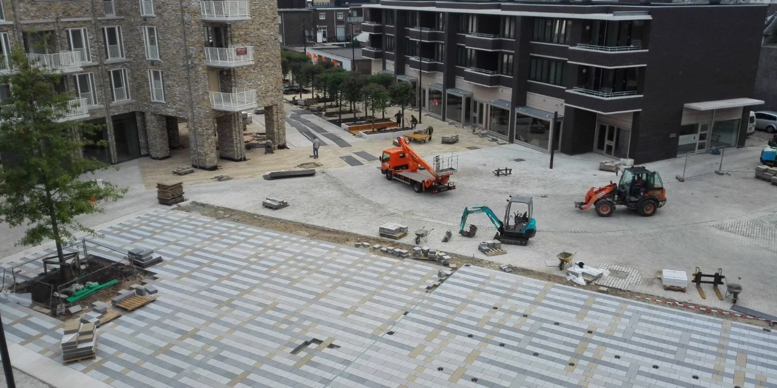 Bouwplaats met werkzaamheden, machines, bouwmaterialen, en een afgeronde tegelvloer naast een bosje en een hoogbouw gebouw.