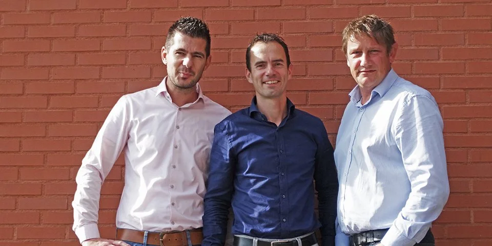 Drie mannen staan voor een rood bakstenen muur, glimlachen, gekleed in nette overhemden.