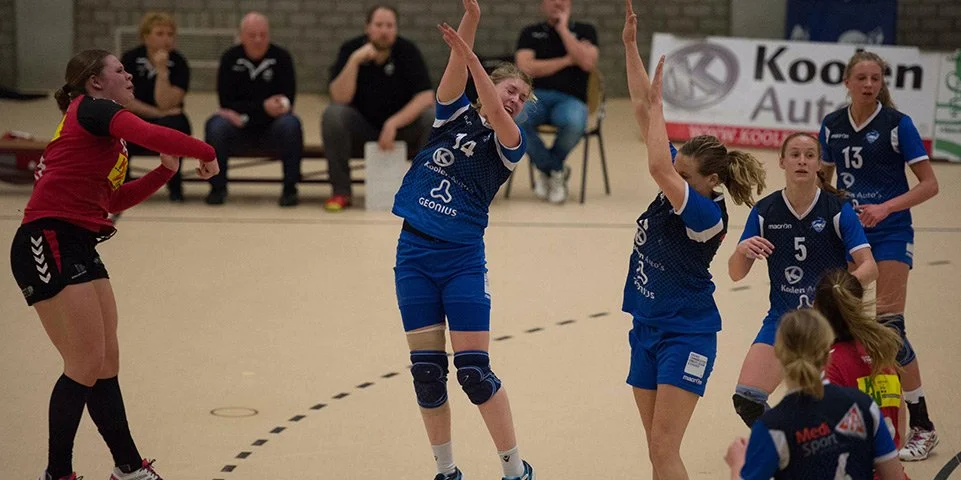 Vrouwelijke handbalsters in blauwe en rode uniforms tijdens een wedstrijd, fans op de achtergrond.