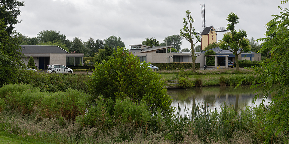 Moderne huizen langs een waterweg met planten en bomen, en een bewolkte lucht.